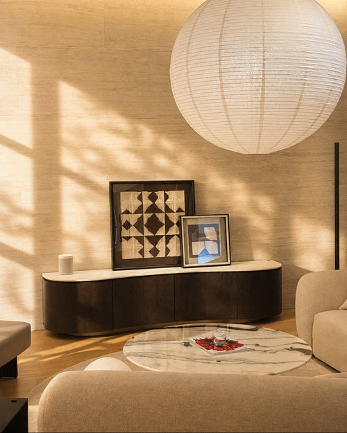 A modern living room scene with a large white spherical paper lantern hanging from the ceiling, a curved dark wood sideboard with framed artwork, a marble coffee table, and beige upholstered seating. Warm sunlight casts shadows across the wall and floor.
