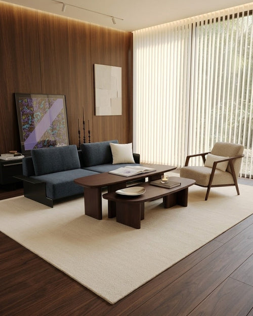 A modern living room featuring a dark blue sectional sofa, a matching dark wood coffee table, a beige armchair, and a large cream-colored area rug on dark wood flooring.