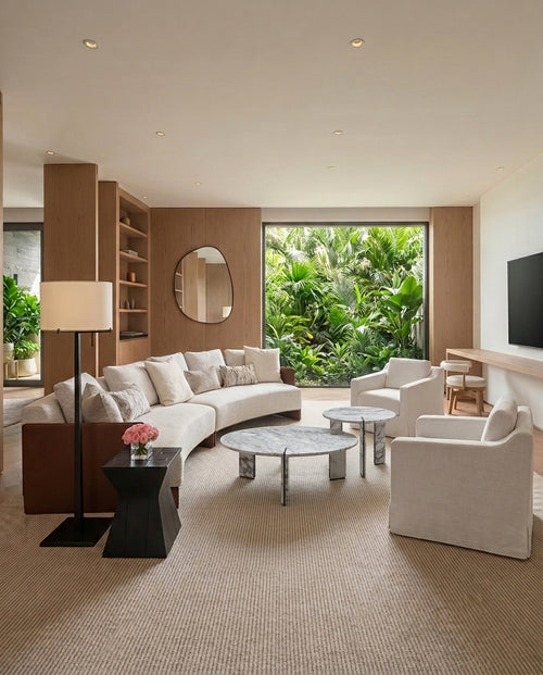 A modern living room with a curved cream sofa, two matching armchairs, round coffee tables, a black side table with a lamp, built-in wood shelving, and a large window overlooking lush green tropical foliage.