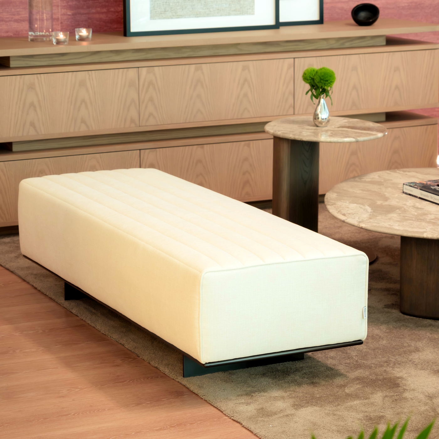 Modern living room with a white ottoman, wooden furniture, and framed pictures on a pink wall.