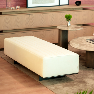 Modern living room with a white ottoman, wooden furniture, and framed pictures on a pink wall.