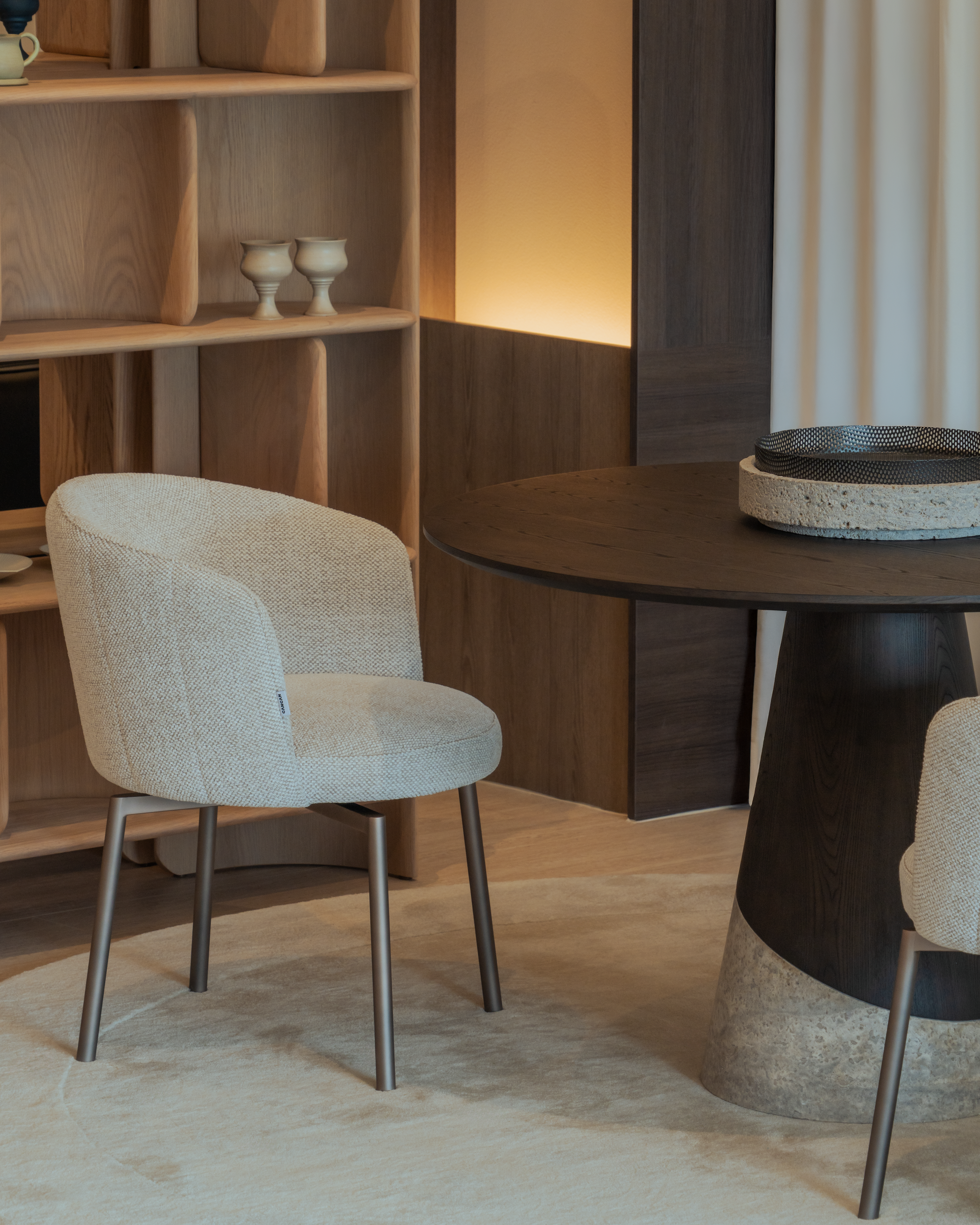 Dining area with a round wooden table and beige chairs against a wooden bookshelf.