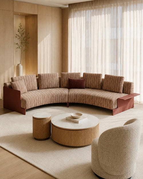A curved upholstered sectional sofa in a warm beige and brown textured fabric with reddish-brown side panels, shown in a styled living room with matching coffee tables, a rug, curtains, and a chair.