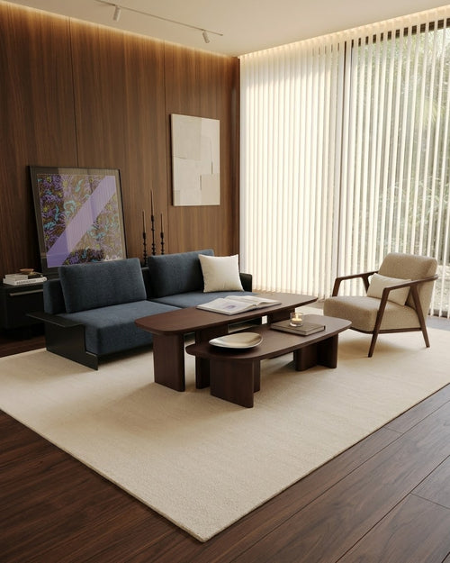 A modern living room setup featuring a dark blue sectional sofa, a matching dark wood coffee table, a beige armchair, and a large cream-colored area rug on dark wood flooring.