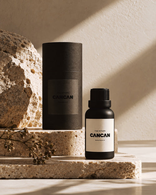 A small black bottle labeled CANCAN next to a tall black cylindrical box, styled on stone blocks with a rock and dried stems in a warm, sunlit setting.