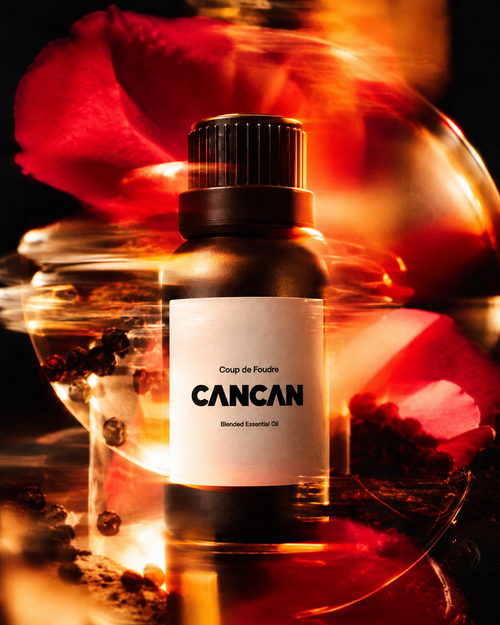 A bottle of CANCAN Coups de Foudre blended essential oil centered against a dark, warm-toned background with red rose petals and glowing amber light effects.