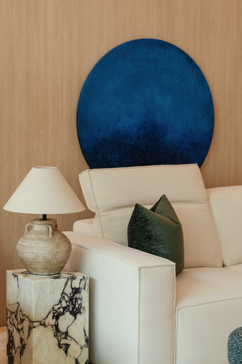 Beige sofa with a green pillow, white lamp on a marble side table, and a blue circular wall art piece.