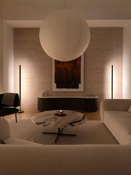 A modern living room interior with a large white spherical pendant light hanging over a round marble coffee table, a cream sofa, floor lamps, and a textured beige wall with framed abstract artwork.