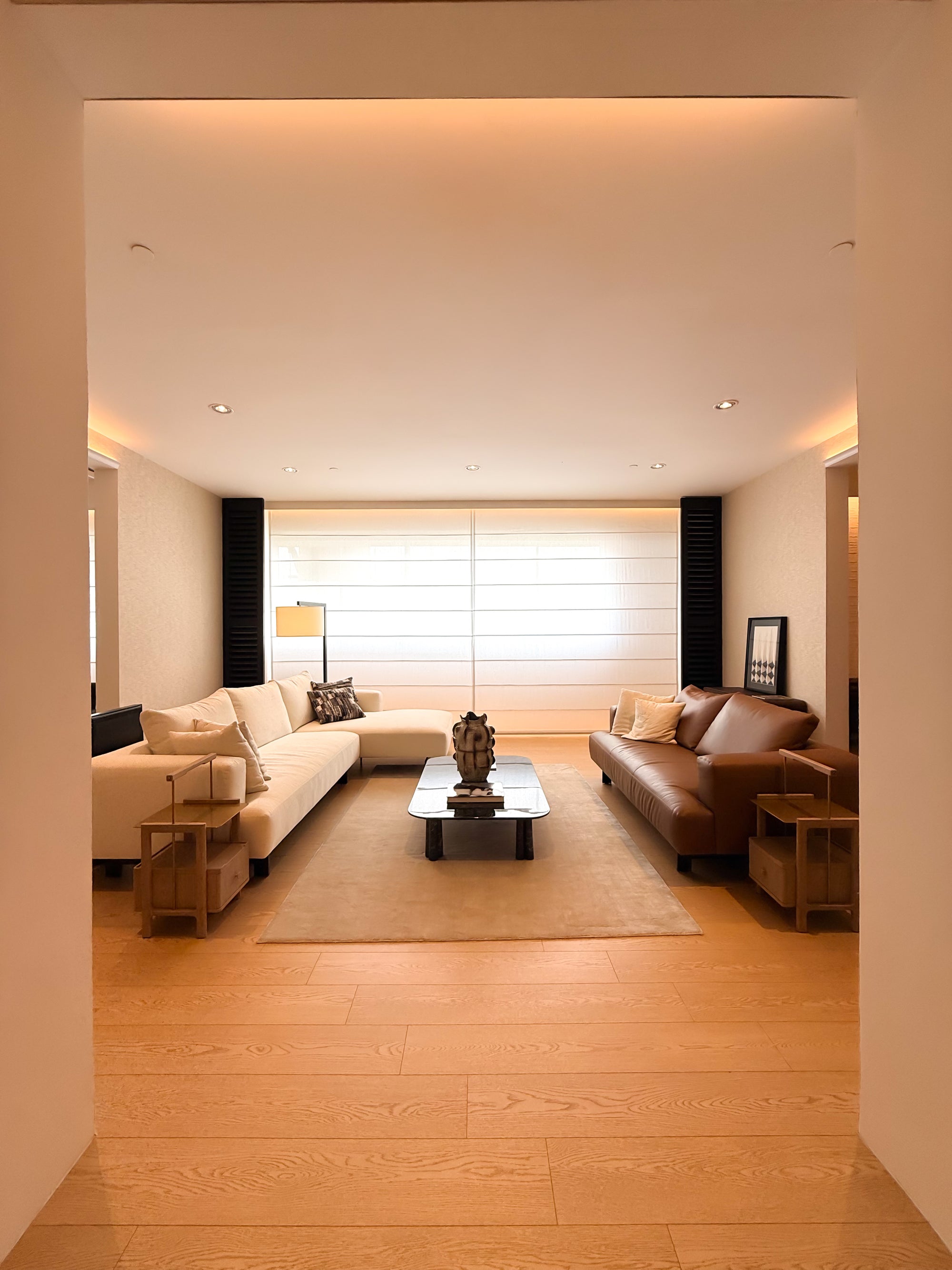 A modern living room with a cream sectional sofa, a brown leather sofa, a low rectangular coffee table, and light wood flooring, viewed from an entryway looking through a doorway.