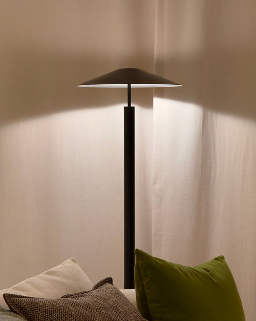 Modern black floor lamp casting light on a wall with pillows on a sofa below.