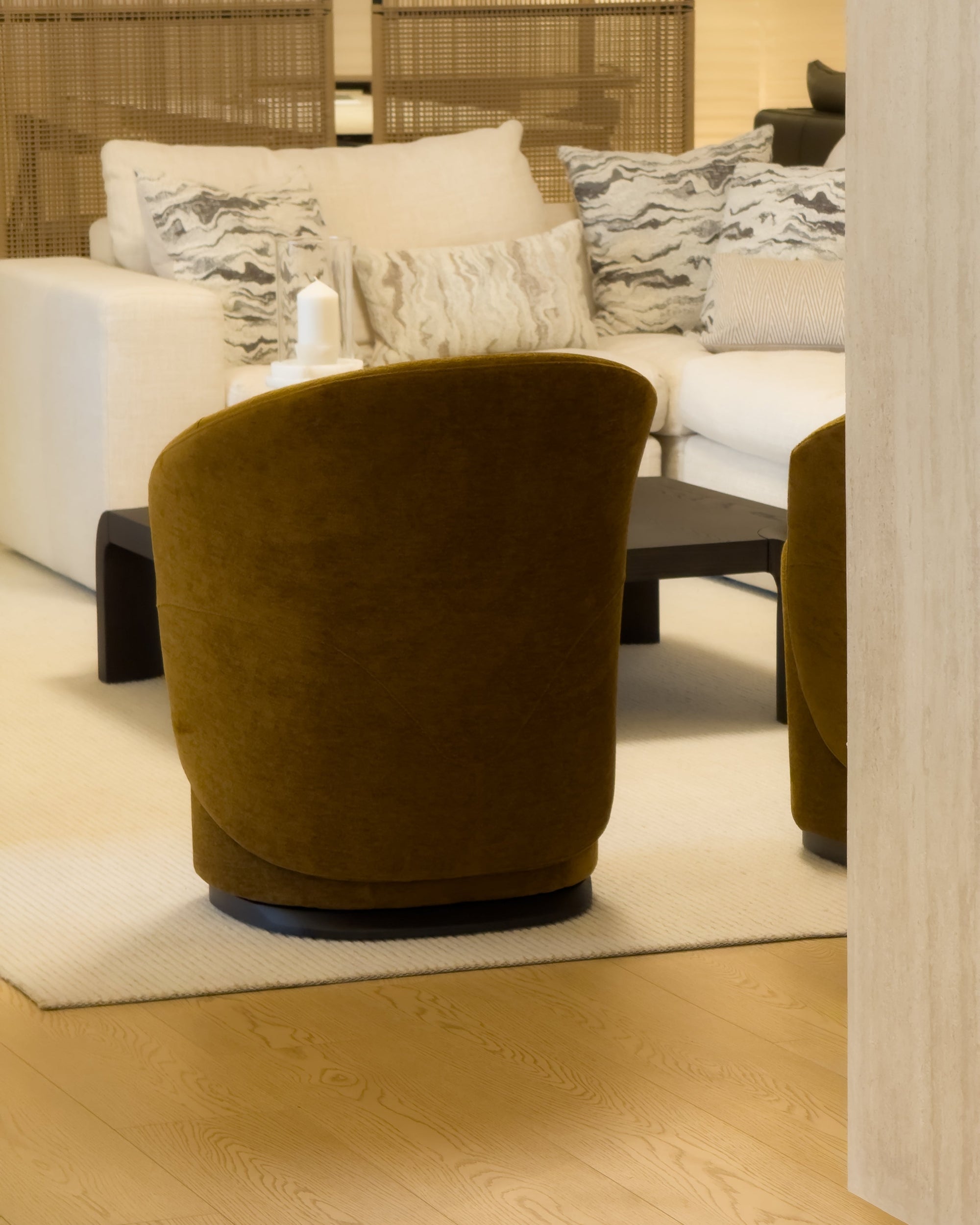 A modern round velvet armchair in a warm olive-brown or mustard-ochre color, positioned in a contemporary living room setting with a white sofa, patterned pillows, a black coffee table, and light wood flooring.