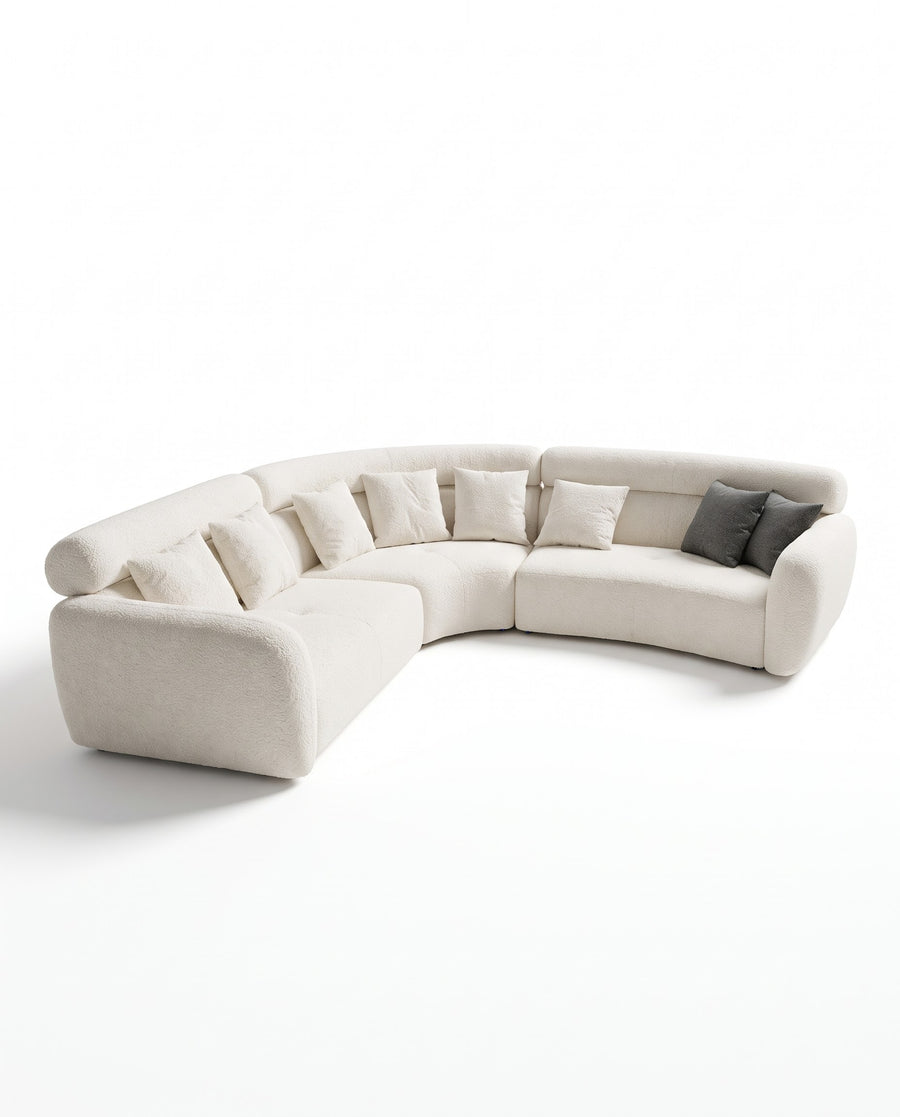 A curved modular upholstered sofa in a light cream fabric, styled with multiple matching throw pillows and two dark gray accent pillows, set against a plain white studio background.