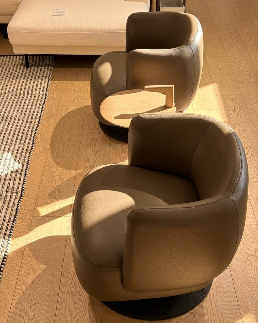 Two modern taupe leather swivel armchairs with small round wooden side tables, set on a light wood floor in a sunlit room.
