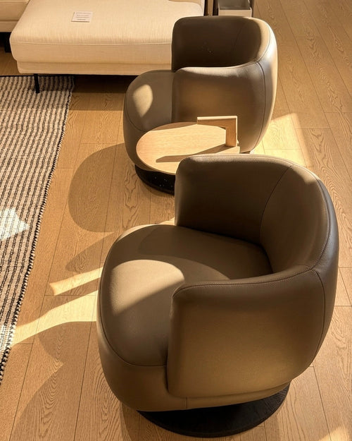 Two modern taupe leather swivel armchairs with small round wooden side tables, set on a light wood floor in a sunlit room.