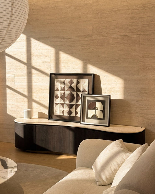 A styled interior scene with two framed abstract geometric wall art prints leaning on a curved dark console table against a warm beige stone wall. Sunlight streams across the wall and floor, and part of a light sofa and hanging lamp are visible.