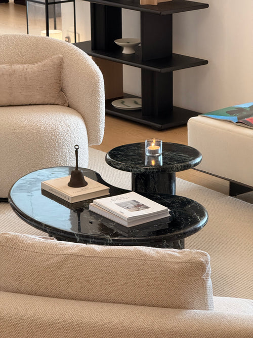 Modern living room scene with black marble nesting coffee tables in the foreground, a cream boucle armchair on the left, and a black shelving unit in the background.