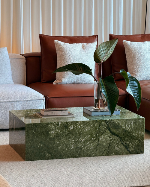 A green marble coffee table in a modern living room setting, styled with a glass vase of green leaves, books, and a brown leather sofa in the background.
