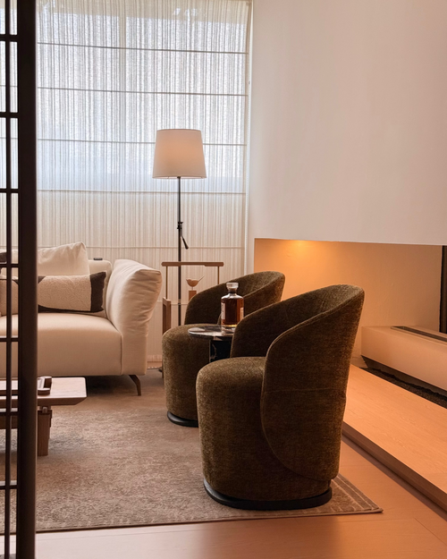 A modern living room scene featuring two dark brown upholstered barrel chairs in the foreground, a cream sofa, a floor lamp, and a warm wood accent wall in the background.