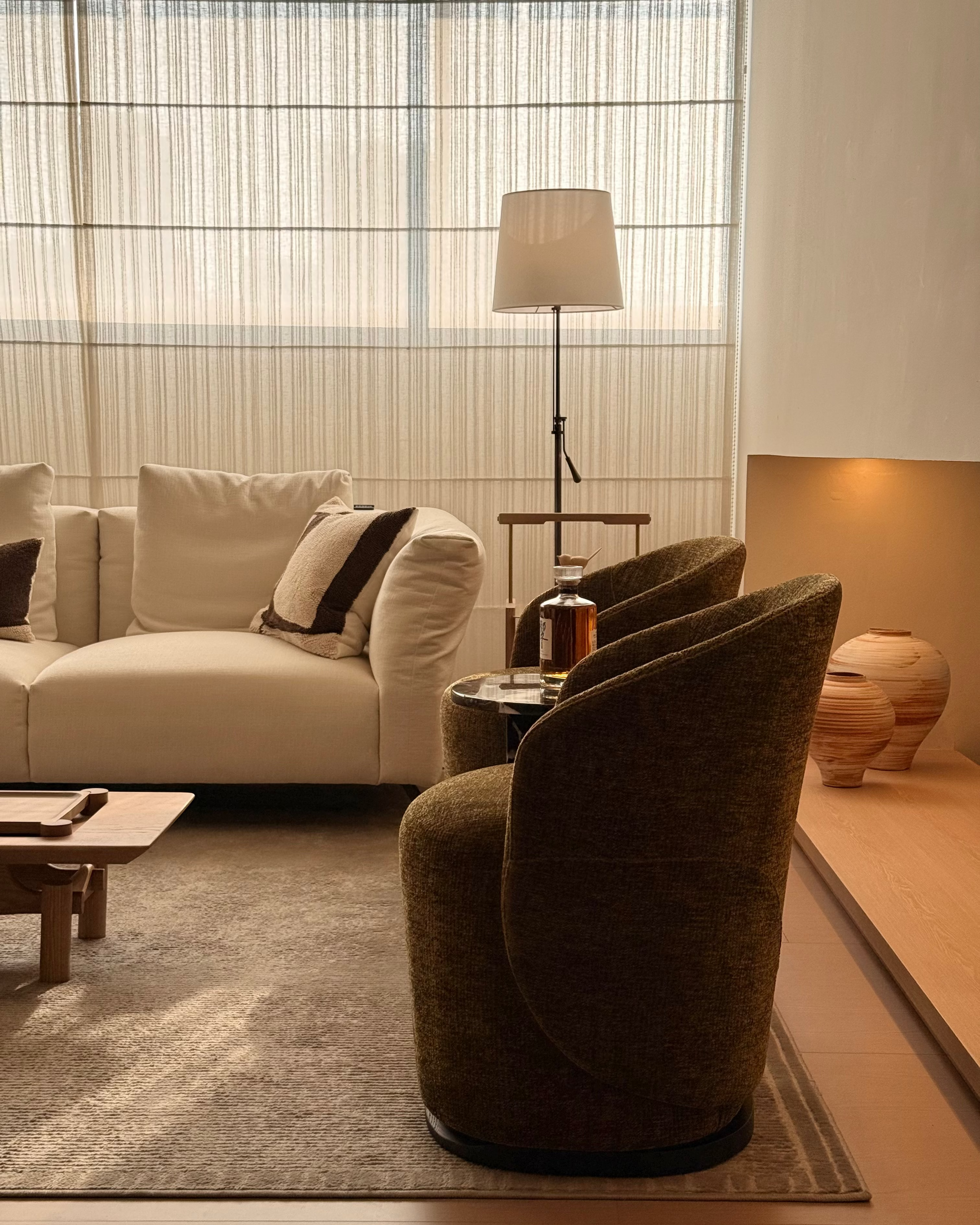 A modern living room scene featuring a prominent dark olive green upholstered accent chair in the foreground, with a cream sofa, a floor lamp, and a textured rug in the background.