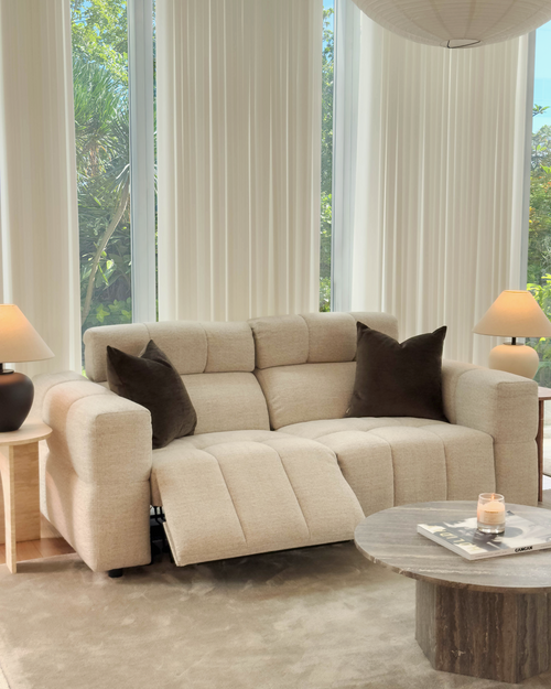 Beige sectional sofa with brown cushions in a living room setting with large windows.