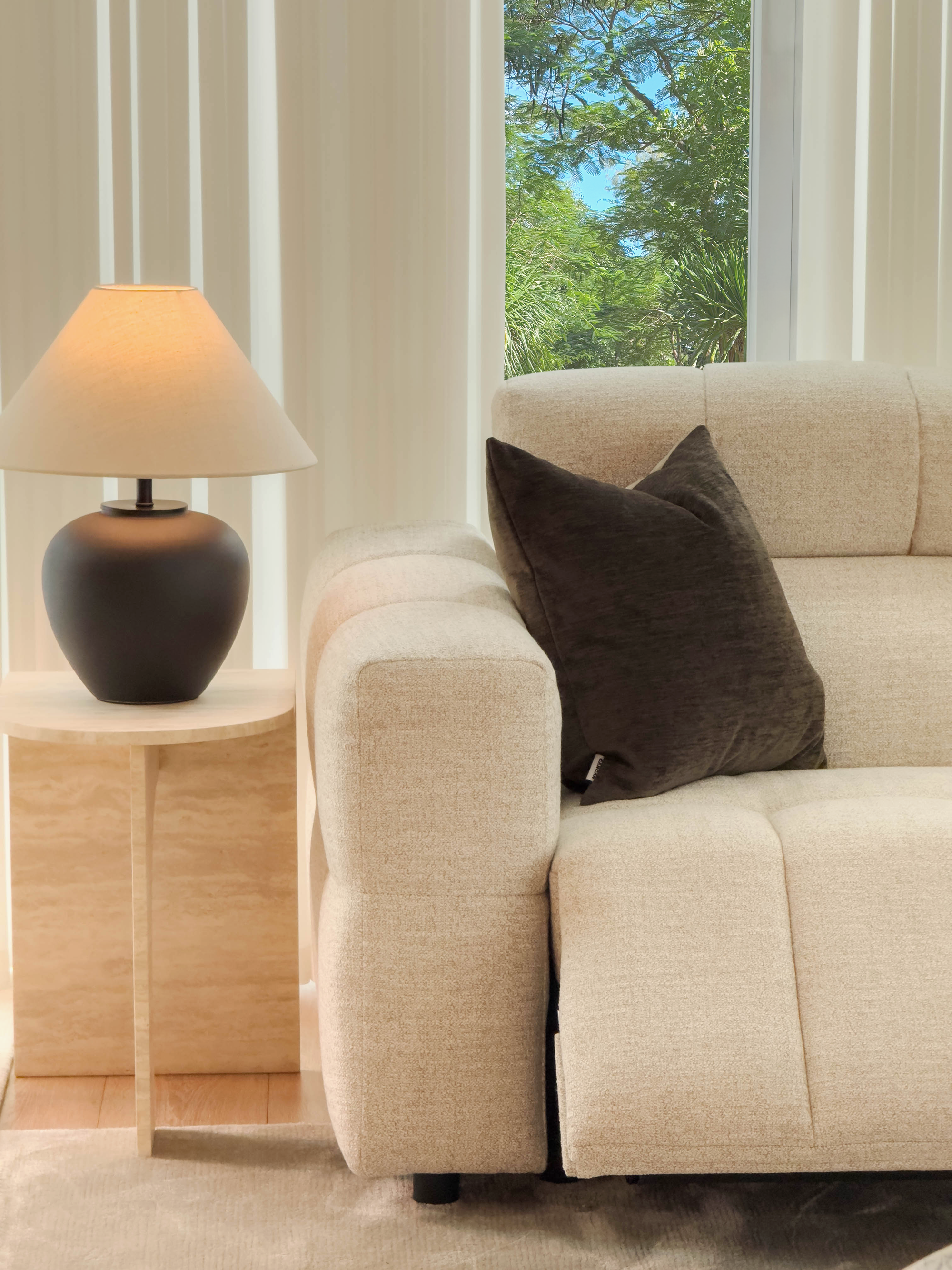 Beige sofa with a brown pillow next to a lamp on a wooden side table, with a window showing greenery in the background.