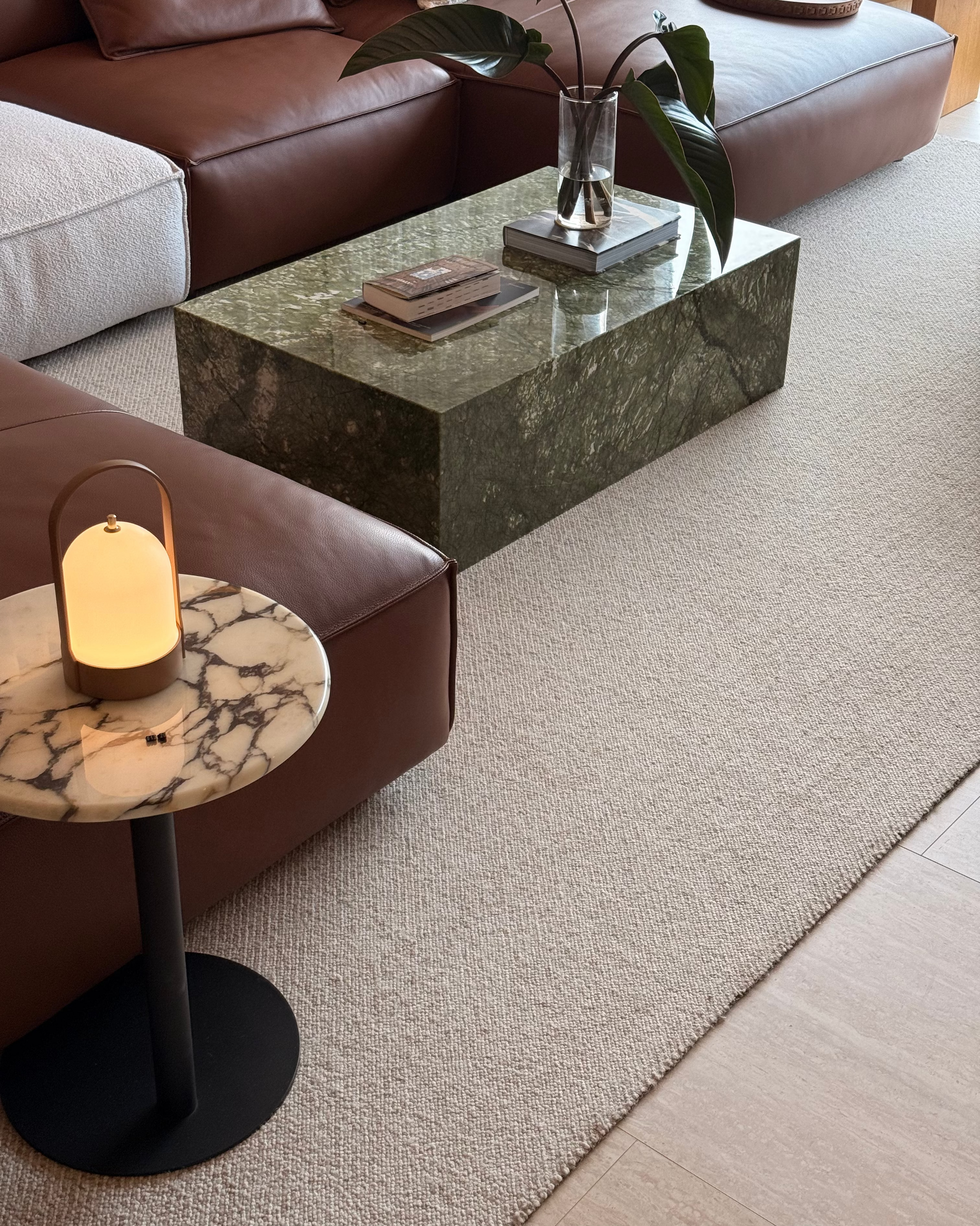 A modern living room scene featuring a dark green marble coffee table, a brown leather sectional sofa, a small round marble side table with a glowing table lamp, and a textured beige rug.
