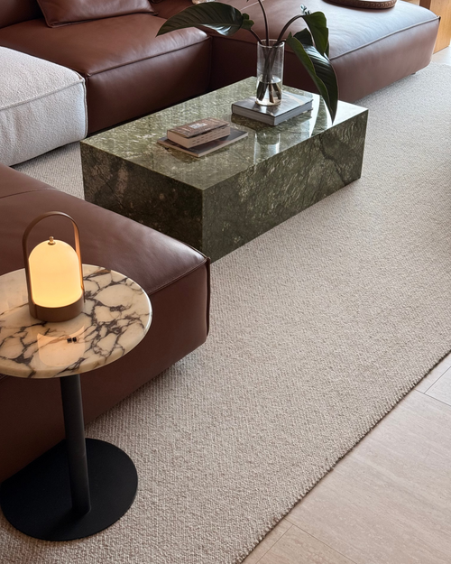 A modern living room scene featuring a dark green marble coffee table, a brown leather sectional sofa, a small round marble side table with a glowing table lamp, and a textured beige rug.