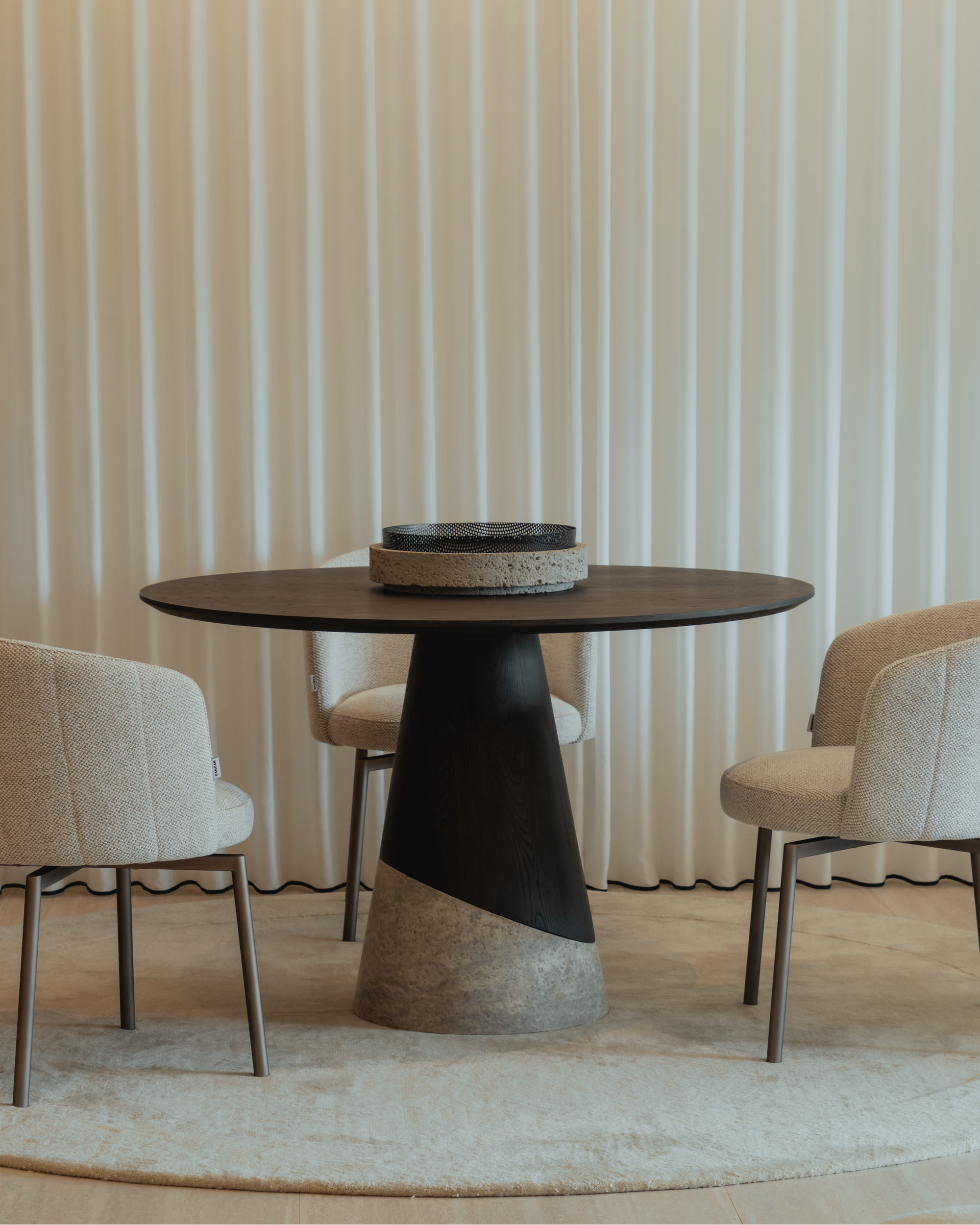 Modern dining table with a black and gray base and beige top, surrounded by chairs against a striped wall.