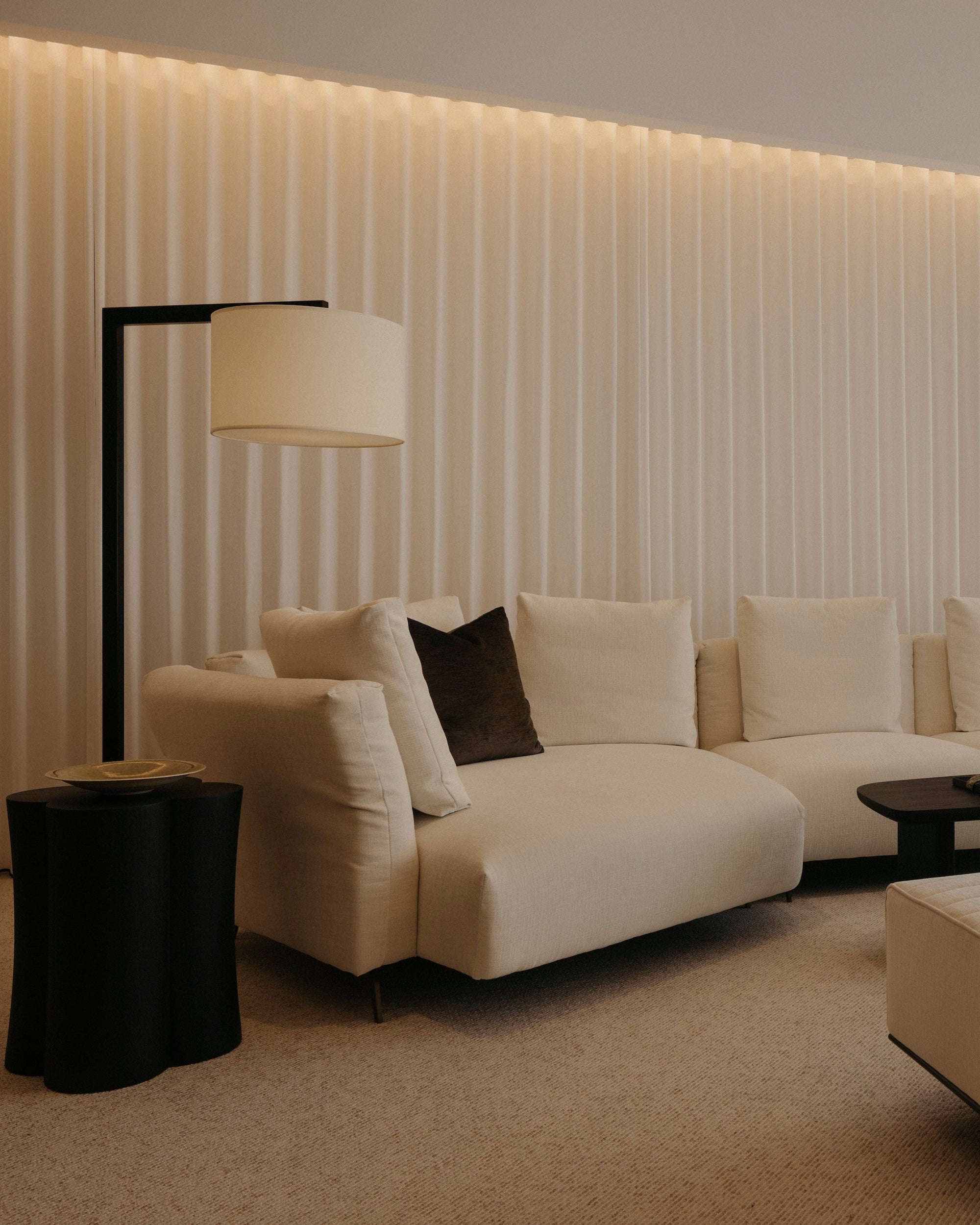 A modern living room featuring a cream sectional sofa with dark accent pillows, a matching cream ottoman, a black floor lamp, and a black coffee table, set against a wall with vertical slats and warm ambient lighting.