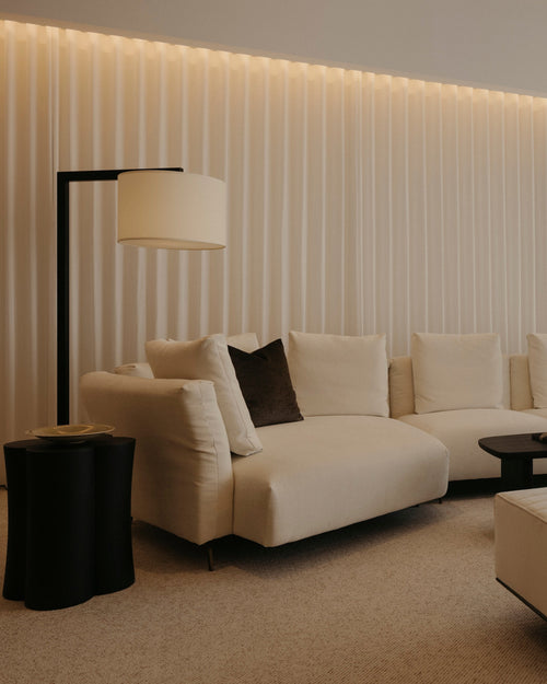 A modern living room featuring a cream sectional sofa with dark accent pillows, a matching cream ottoman, a black floor lamp, and a black coffee table, set against a wall with vertical slats and warm ambient lighting.