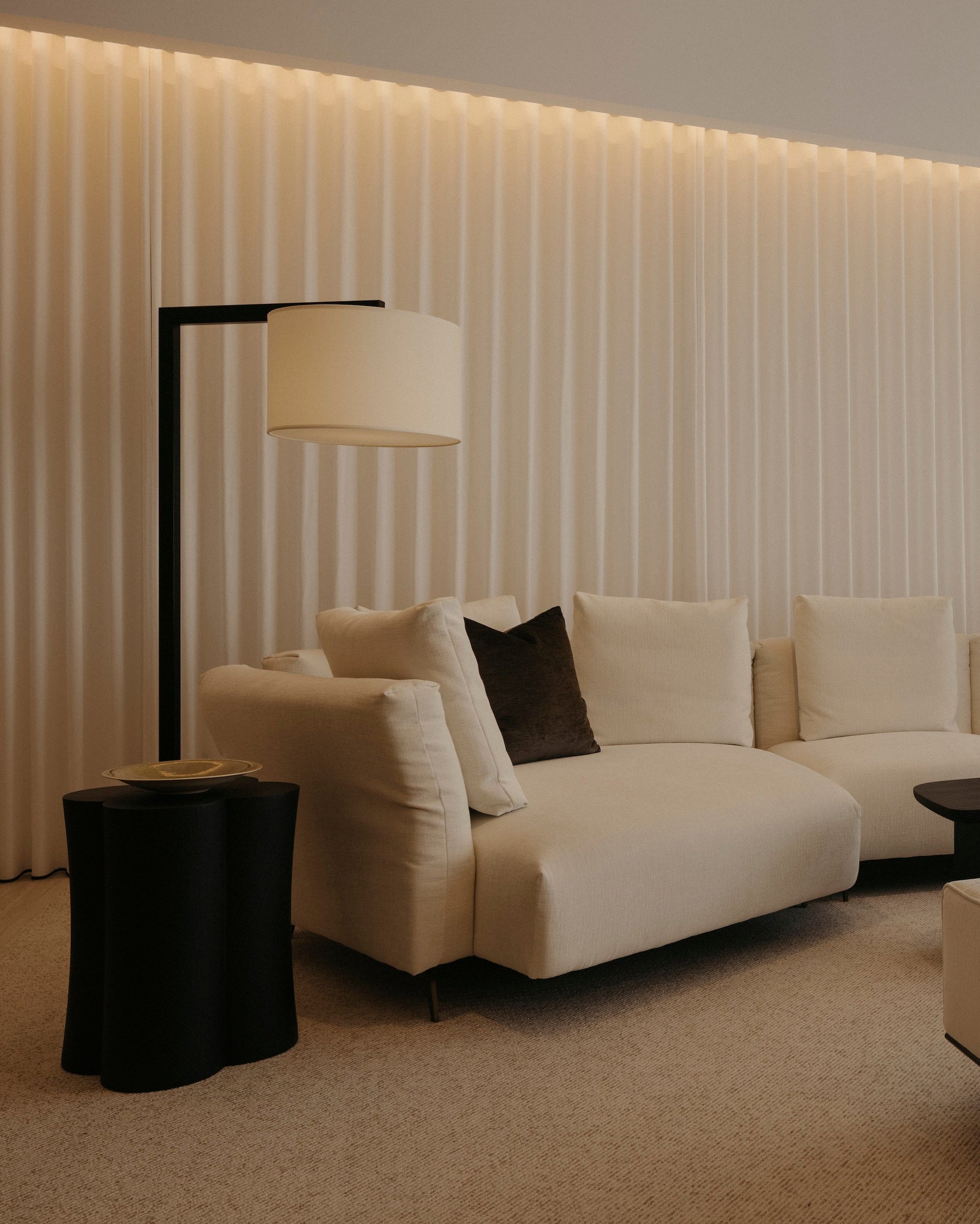 A modern living room featuring a cream sectional sofa with dark accent pillows, a matching cream ottoman, a black floor lamp, and a black coffee table, set against a wall with vertical slats and warm ambient lighting.
