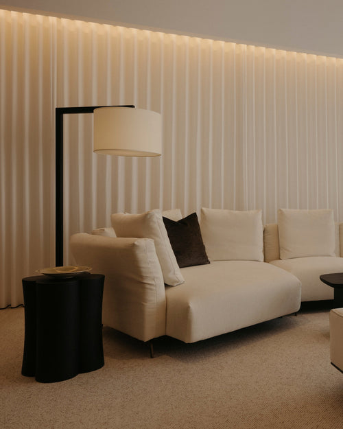 A modern living room featuring a cream sectional sofa with dark accent pillows, a matching cream ottoman, a black floor lamp, and a black coffee table, set against a wall with vertical slats and warm ambient lighting.