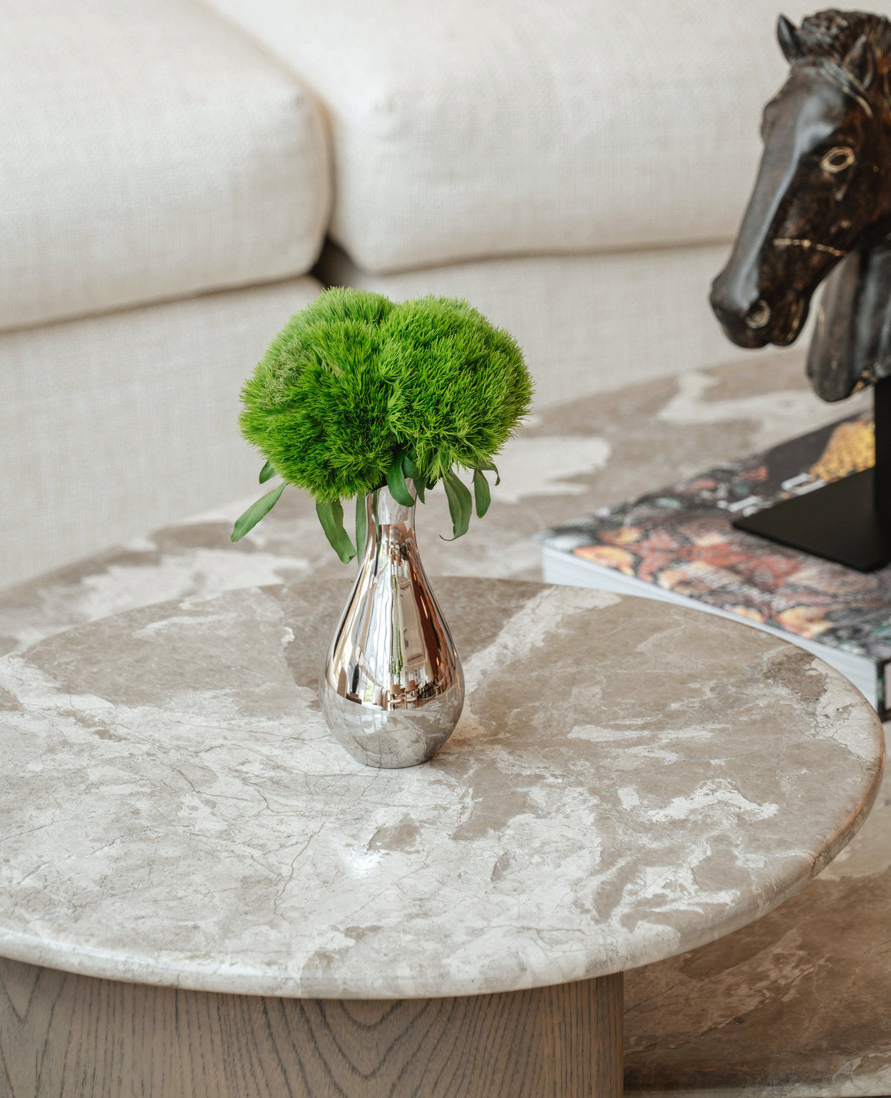 Round marble coffee table with a silver vase containing green foliage, next to a beige sofa and decorative horse figure.