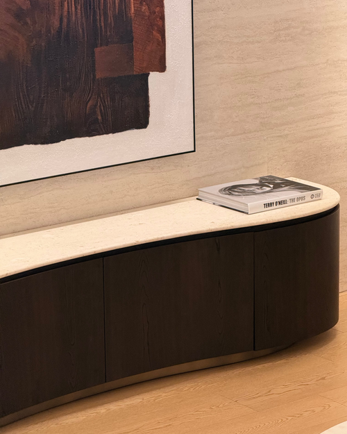 A curved modern media console with dark wood cabinets and a light stone top, styled in a contemporary room setting with a book on top.