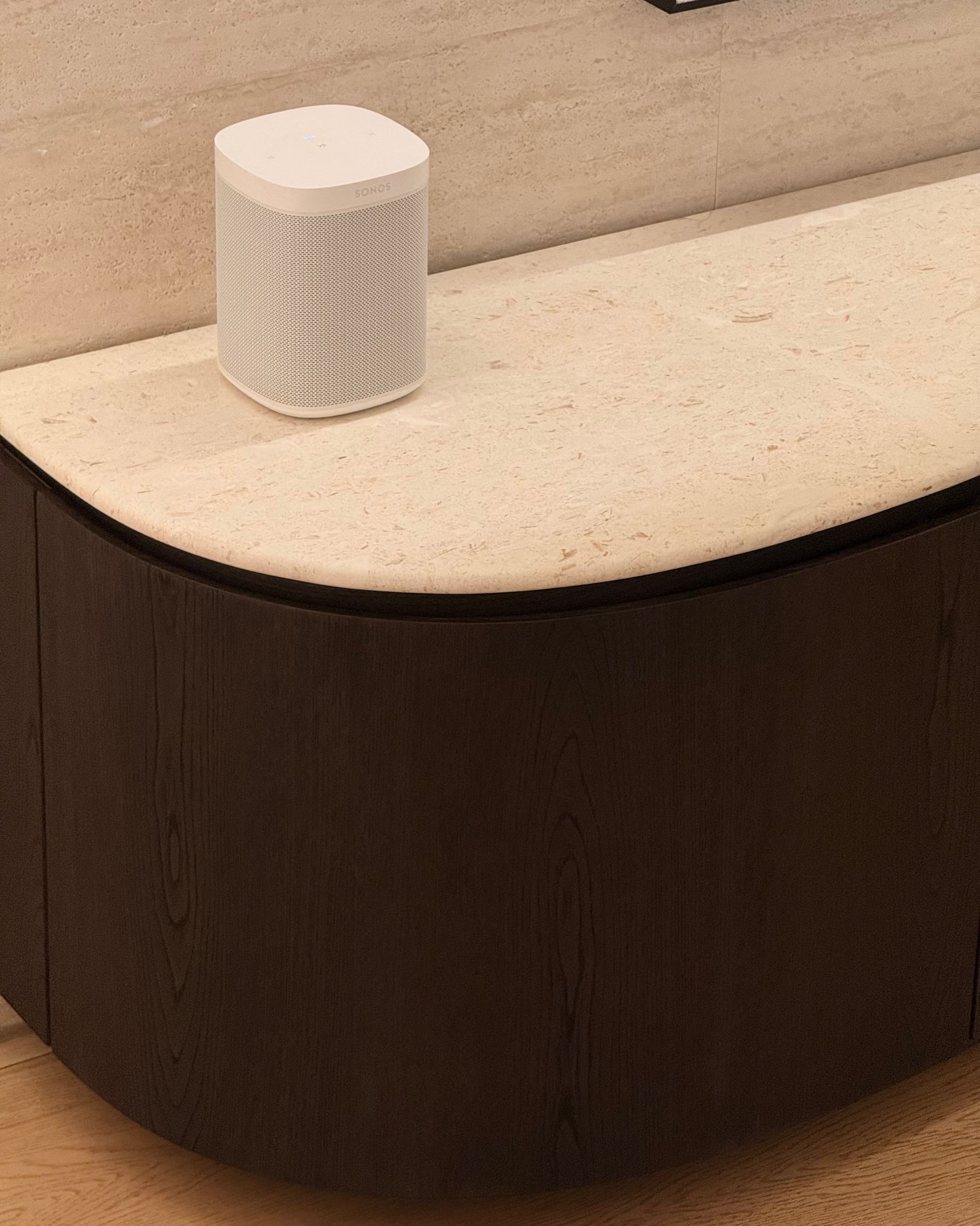 A white cylindrical smart speaker with rounded corners sits on a curved cream stone surface above a dark wood cabinet in a modern interior setting.