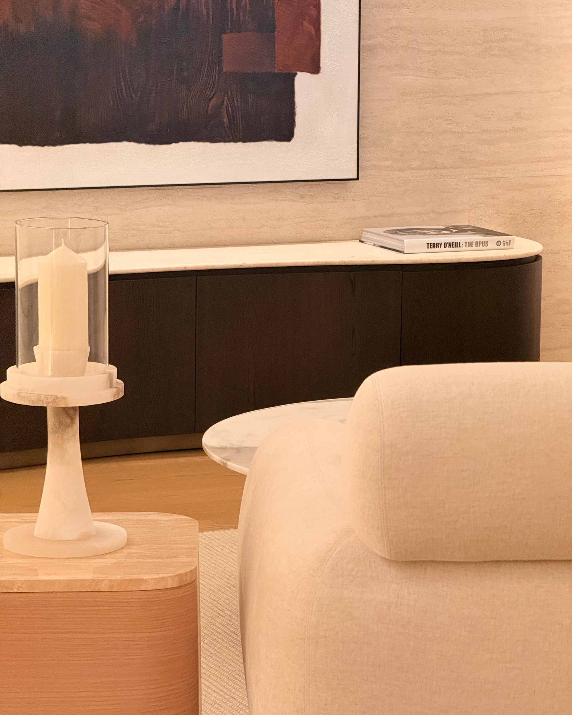 A close-up lifestyle shot of a modern living room corner featuring a beige upholstered armchair, a marble candle holder on a wooden side table, and a curved dark wood and cream media console against a textured beige wall.