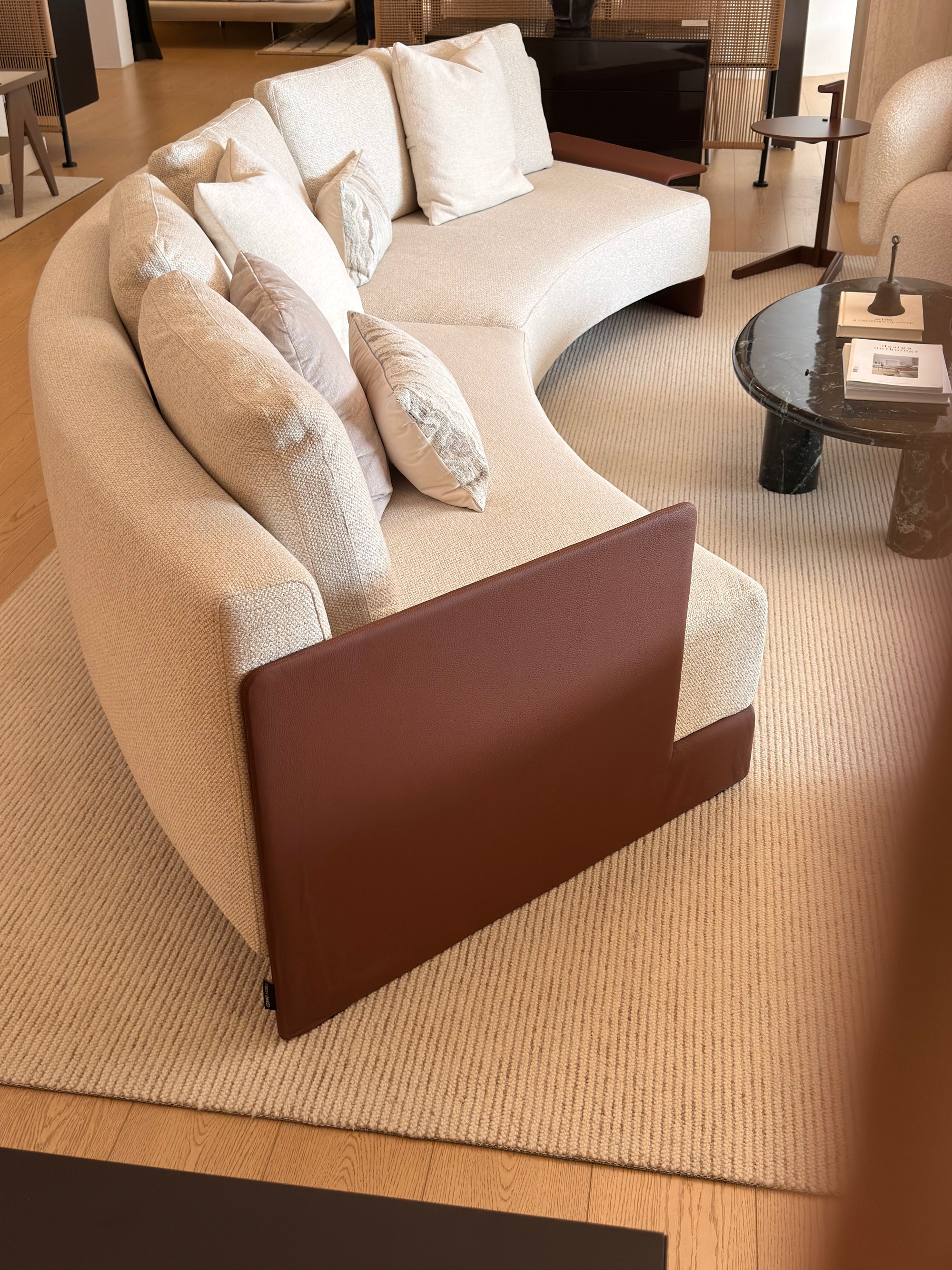 A curved upholstered sofa in a warm cream or beige fabric with a contrasting dark brown leather front panel, styled in a modern living room setting on a textured rug.