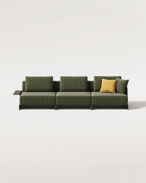 A modern low-profile modular sofa upholstered in textured olive green fabric, featuring a small dark side table on the left and a contrasting yellow accent pillow, set against a plain light gray background.